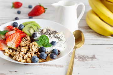 White ceramic bowl of homemade granola with yogurt, fresh berries and fruits on white wooden table background.