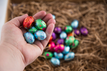 Human hand holding a selection of small colorful foil wrapped eggs. A pile are underneath lying on paper packing straw, out of focus. Shows scale and size of mini eggs. Caucasian hand, male or female.