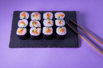 Maki sushi with salmon and avocado on a slate plate with chopsticks on purple background. Japanese food.