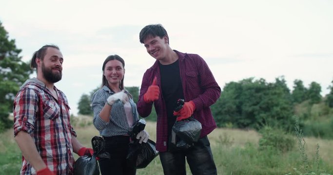 Team of volunteers with garbage bags talk, show thumbs and smile on nature.