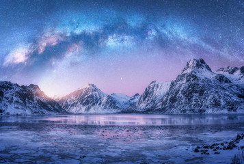 Milchstraße über gefrorener Meeresküste und schneebedeckten Bergen im Winter nachts auf den Lofoten-Inseln, Norwegen. Arktische Landschaft mit blauem Sternenhimmel, Wasser, Eis, schneebedeckten Felsen, Milchstraße. Weltraum und Galaxie