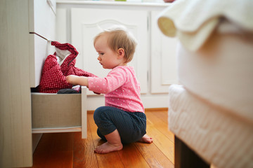 Adorable toddler girl at home, opening the drawer of dresser and taking out clothes