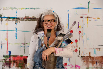 smiling older woman, proud artist, in her fifties with grey hair and glasses and many paintbrushes