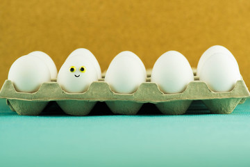 Happy smiling easter egg among simple white eggs in a paper tray on turquoise and mustard yellow bacground. Funny easter scene.