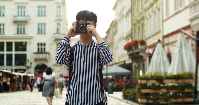 Cheerful Asian young man traveller with backpack taking photo with photocamera and smiling in center city of european town. Happy Chinese guy student tourist having sightseeing tour. Traveling concept