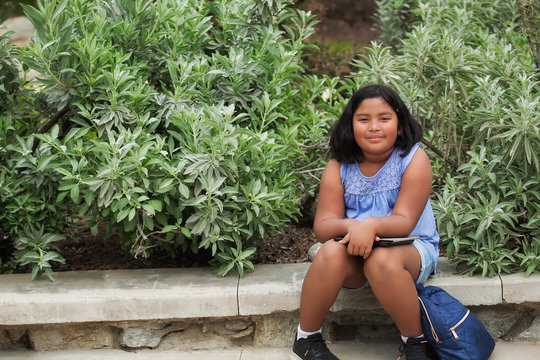 An elementary school girl who is sitting down while she waits with her tablet and school backpack, surrounded by green plants.