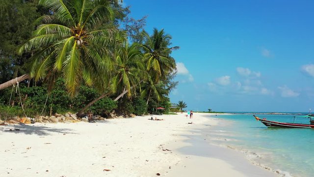 Exotic beach with white sand under shadow of palm trees with green leaves and coconut, anchored boat on tropical shore, Thailand