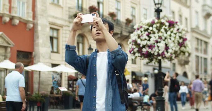 Handsome Asian young man traveller taking photos with phone camera in center city of european town while walking the street. Chinese guy student tourist having sightseeing tour. Traveling concept.