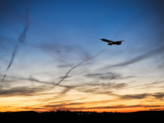 Fototapeta na wymiar Dark silhouette of passenger plane with landing gear still down ascending into partially cloudy sky with soft orange red urban skyline in background.
