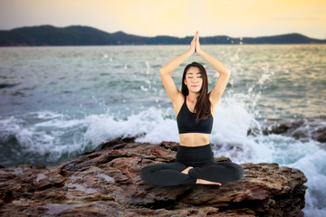 Fototapeta na wymiar Attractive smiling woman practicing yoga on the rocks against the beach background At sunset