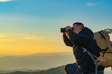 The young man with a lifted backpack looked in binoculars sitting on top of the mountain, enjoying...