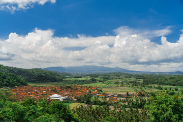 Traditional Balinese village in the valley of the mountains. Orange roofs of houses, coconut palms and rice fields against the background of mountains and clouds.