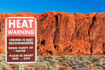 Heat warning sign informs travelers of the dangers of heat exhaustion from hiking in the desert climate. Blurred scenic red sandstone formations and desert scrub landscape background. Blue sky
