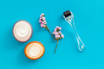 Cosmetology and skin care concept. Dermaroller, creams near flowers on blue background top-down