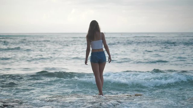 Girl in walking to denim shorts sea on stone beach. Waves crashing against shore. 4k