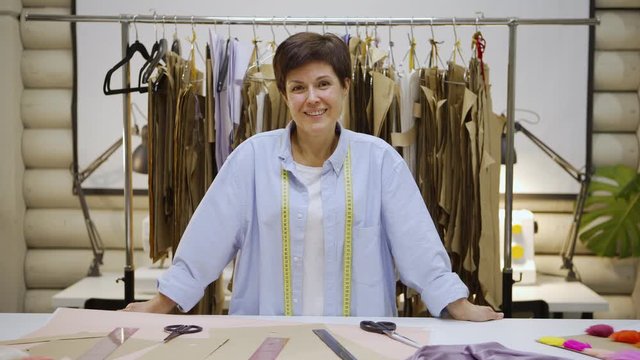 Tilt up portrait of smiling female tailor with short hair looking at camera standing at table in dressmaking studio, rail with sewing patterns in background