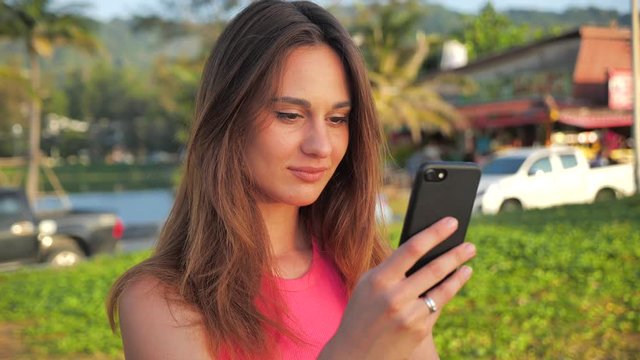 Close up portrait of young woman in a pink short shirt. She stands in the site palms in the background. Female used smartphone. 4k