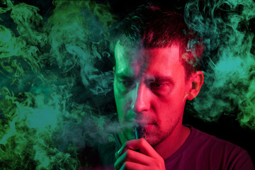 A man holds in his hand and drags on an electronic cigarette with a vape while smoking and releases smoke to the sides, his face is highlighted with green neon chemical light, and head is in clouds