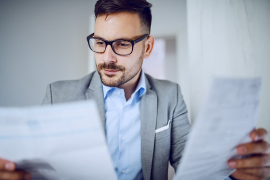 Head shot of hardworking sophisticated caucasian businessman in suit and with eyeglasses looking at documents while sitting in his office.