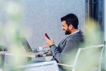 Young charming Caucasian bearded businessman sitting on terrace of cafe and responding to message...