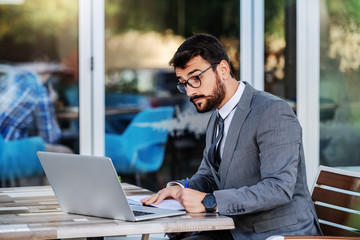 Young dedicated Caucasian elegant bearded businessman in suit with eyeglasses sitting at terrace of his favorite cafe, taking notes in agenda and looking at laptop.