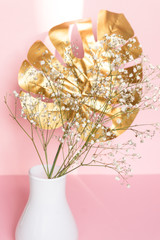 White Vase with golden tropical leaf and gypsophila flowers on table against pink background