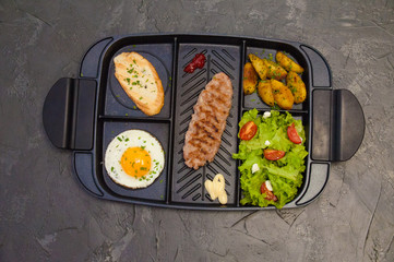 Breakfast, fried steak or cutlet, salad with tomato and green vegetables, toast, potatoes, egg on a black plate. Concept of a healthy diet, diet, menu, good nutrition.