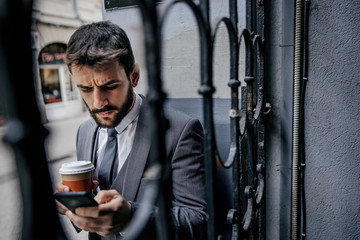 Young bearded businessman standing next to an old iron fence in font of building, holding cup with fresh morning coffee and typing message on smart phone. - Powered by Adobe