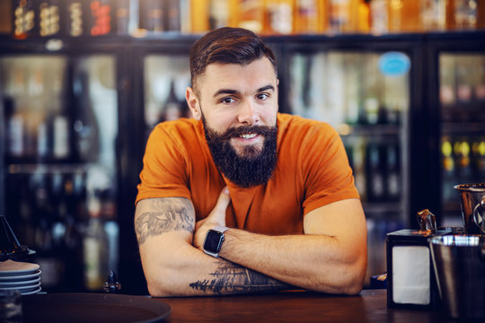Portrait of handsome bearded smiling positive tattooed barman leaning on bar counter, looking at camera and waiting for customers to order drinks.