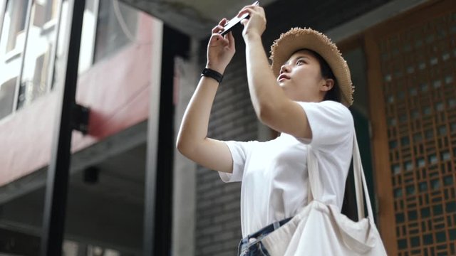 Beautiful asian woman wearing a fedora hat using a smartphone taking a photo while standing a small street in the old town. Happy holiday travel lifestyle teenager.