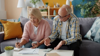 Senior man and woman are counting money looking through bills busy with finance on couch at home. Payments, family life and apartment concept. - Powered by Adobe