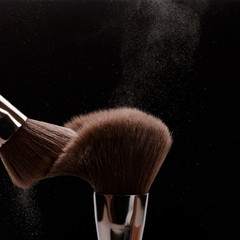Make-up brush with beige powder explosion on black background. Creative splash. Professional powder. Cosmetics brush releasing a cloud of sparkling face powder. Copy space. Beauty and makeup concept