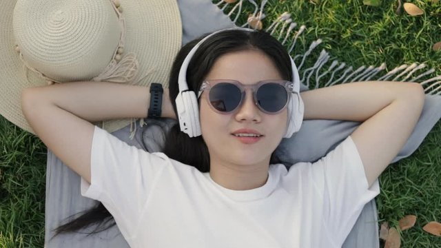 Beautiful young asian woman wearing sunglasses and headphones while lying on the floor grass outdoors at a public park on the beautiful sunset. Sleeping on the grass feeling relaxed in vacation summer