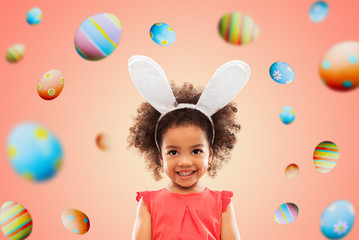 Obraz na płótnie Canvas easter, holidays and childhood concept - happy little african american girl wearing bunny ears headband over living coral background and colored eggs