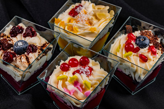 Cropped image of four trapezoid glasses with individual mixed desserts on isolated dark background
