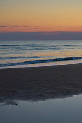 Obraz premium Colorful sunset at the tropical beach, sun behind clouds reflects on water and waves with foam hitting sand.