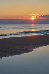 Colorful sunset at the tropical beach, sun behind clouds reflects on water and waves with foam hitting sand.