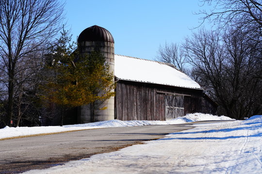 Old vintage wooden abandon rustic barn and grain mill covered in snow from the Winter Season sit near walking path of Riverside Park in St. Cloud, Wisconsin - Powered by Adobe