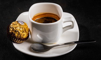 Close-up of black coffee cup on white saucer next to teaspoon and candy in foil wrapper, high angle