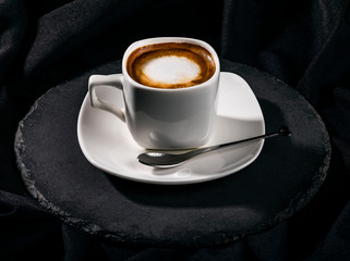 One white ceramic cup of coffee with milk on saucer on round board, isolated black background