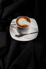 White ceramic cup of coffee with milk on plate next to teaspoon, isolated black fabric background