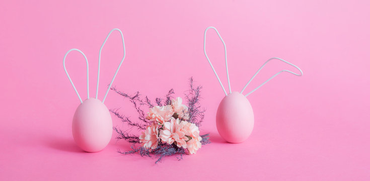 Two Easter pink eggs with rabbit ears made of wire and a bouquet of flowers between them on a pink background. The concept of a happy Easter. Gentle postcard in calm colors, a place to copy.