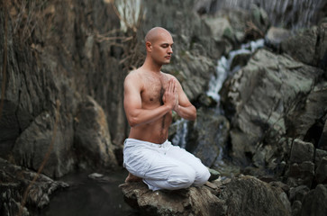 Buddhist. Bald man. Meditation in the mountains against the backdrop of a waterfall. Prayer.