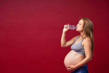 A pregnant woman drinks water on a red background. Thirsty. Enjoys the taste of pure water....