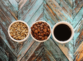 Green raw beans, roasted coffee beans and black coffee in three white cups on turquoise wooden background top view
