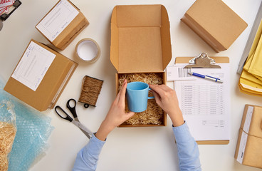 delivery, mail service, people and shipment concept - female hands packing mug to parcel box with...