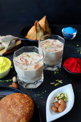 Assorted Indian Mahashivratri or Holi festival food like samosa, kachori, Kaju Katli, Puran Poli, dry fruits & Almond milk Thandai drink with Happy Holi wishes over black background with copy space.