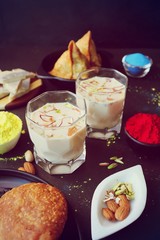 Assorted Indian Mahashivratri or Holi festival food like samosa, kachori, Kaju Katli, Puran Poli, dry fruits & Almond milk Thandai drink with Happy Holi wishes over black background with copy space.
