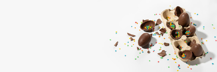 Broken Easter Chocolate eggs in a tray and multi-colored decorations on a light background. Easter concept, easter treats,. Banner. Flat lay, top view