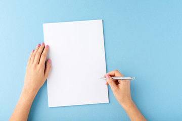 Overhead shot of female hands writing with pen over empty white sheet of paper on blue background